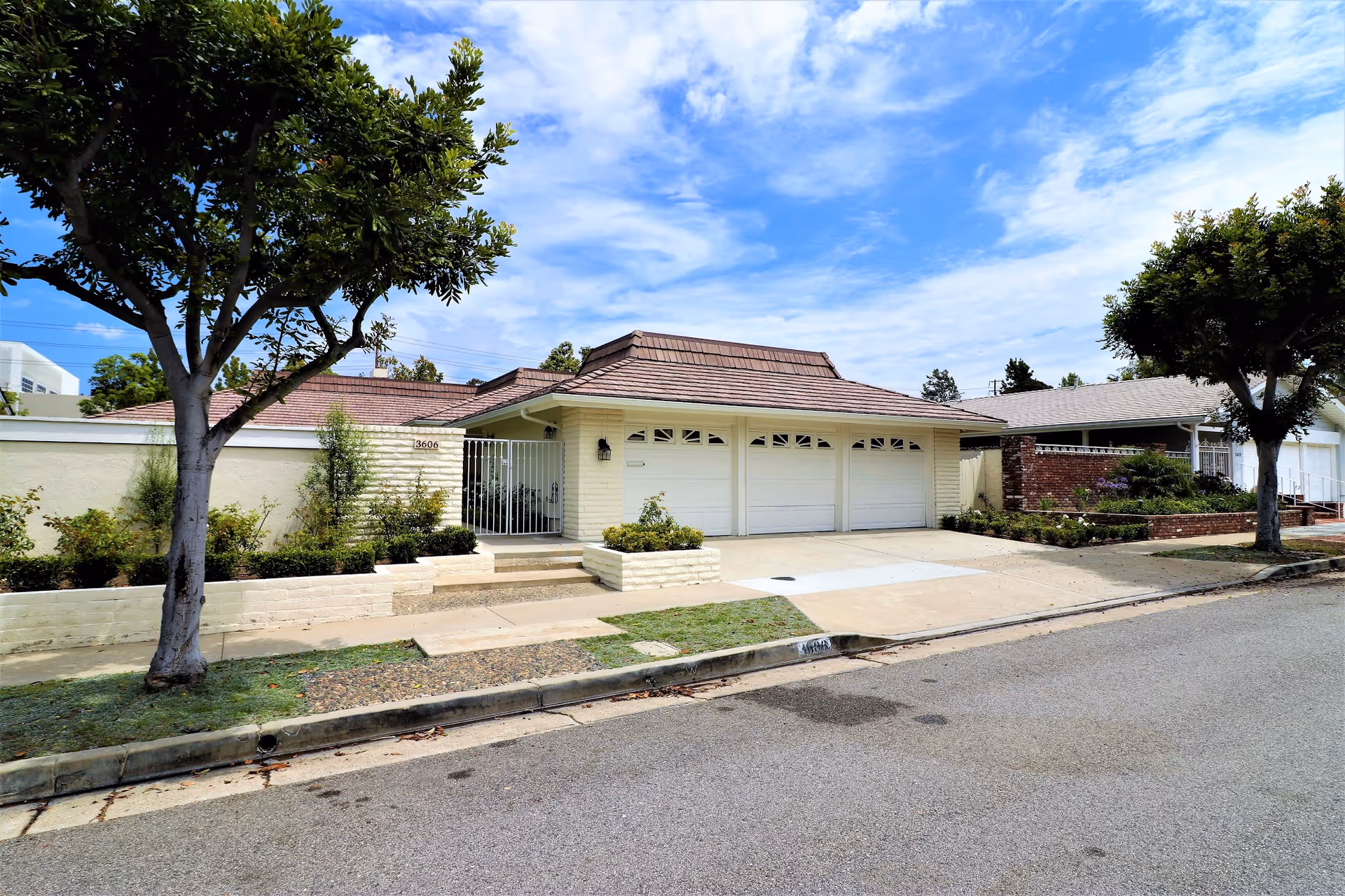 Exterior view of a single-story residential building with a three-car garage, a tiled roof, and a small garden area with shrubs and trees in front. The sky is partly cloudy with blue patches visible.