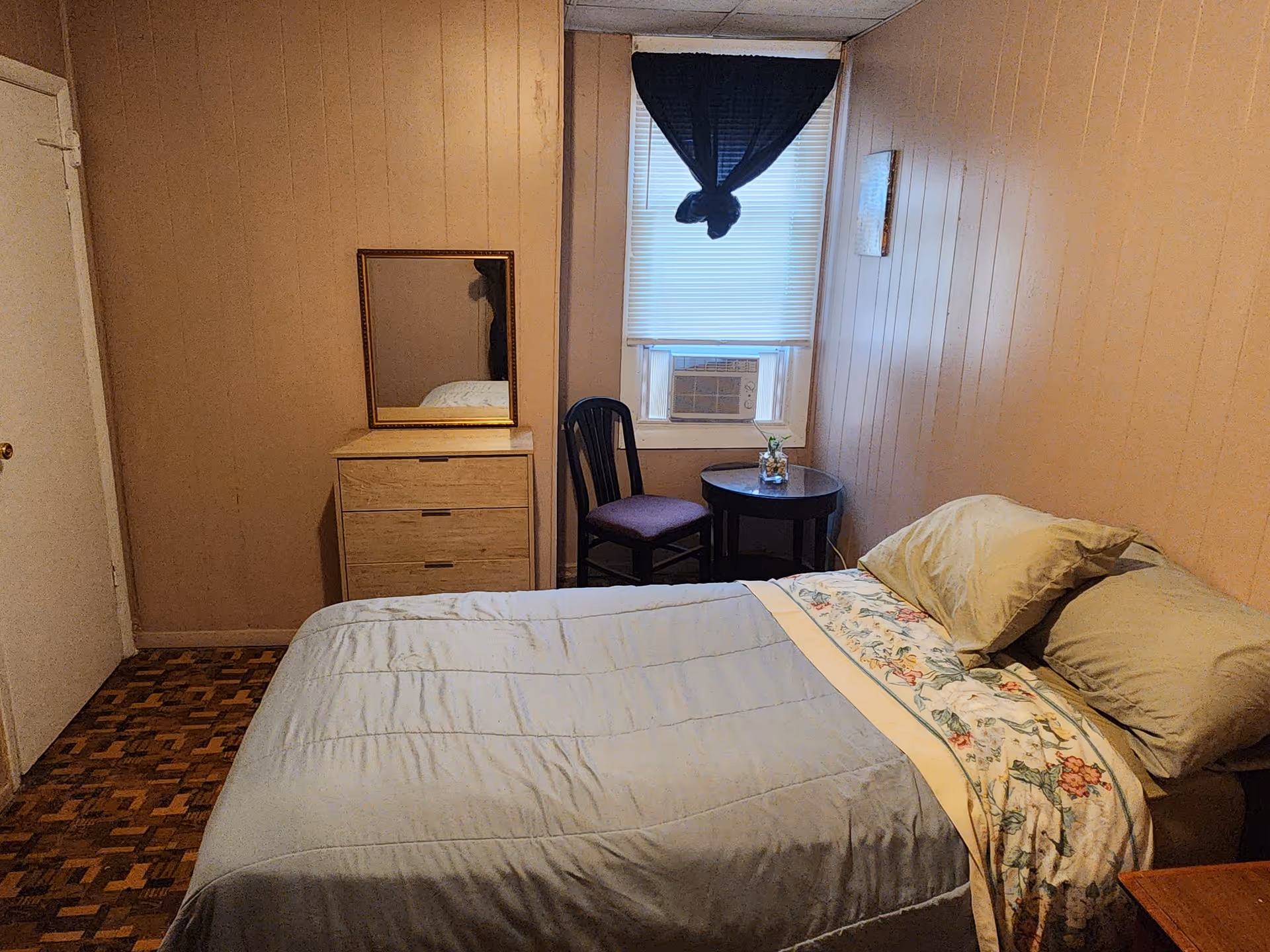 Small bedroom in a senior living facility with bed, dresser, window, and seating.