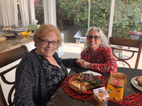 Two women smiling at a dining table with plates of food and a view of a garden through sliding glass doors.