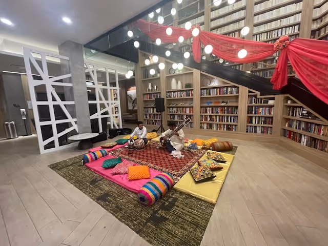 A cozy library lounge with floor cushions and two people playing music near a red-draped staircase.