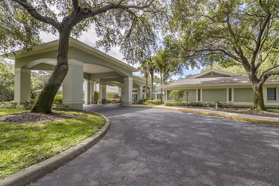 Front entrance with a covered porte-cochère and driveway at a single-story building surrounded by trees and landscaping.