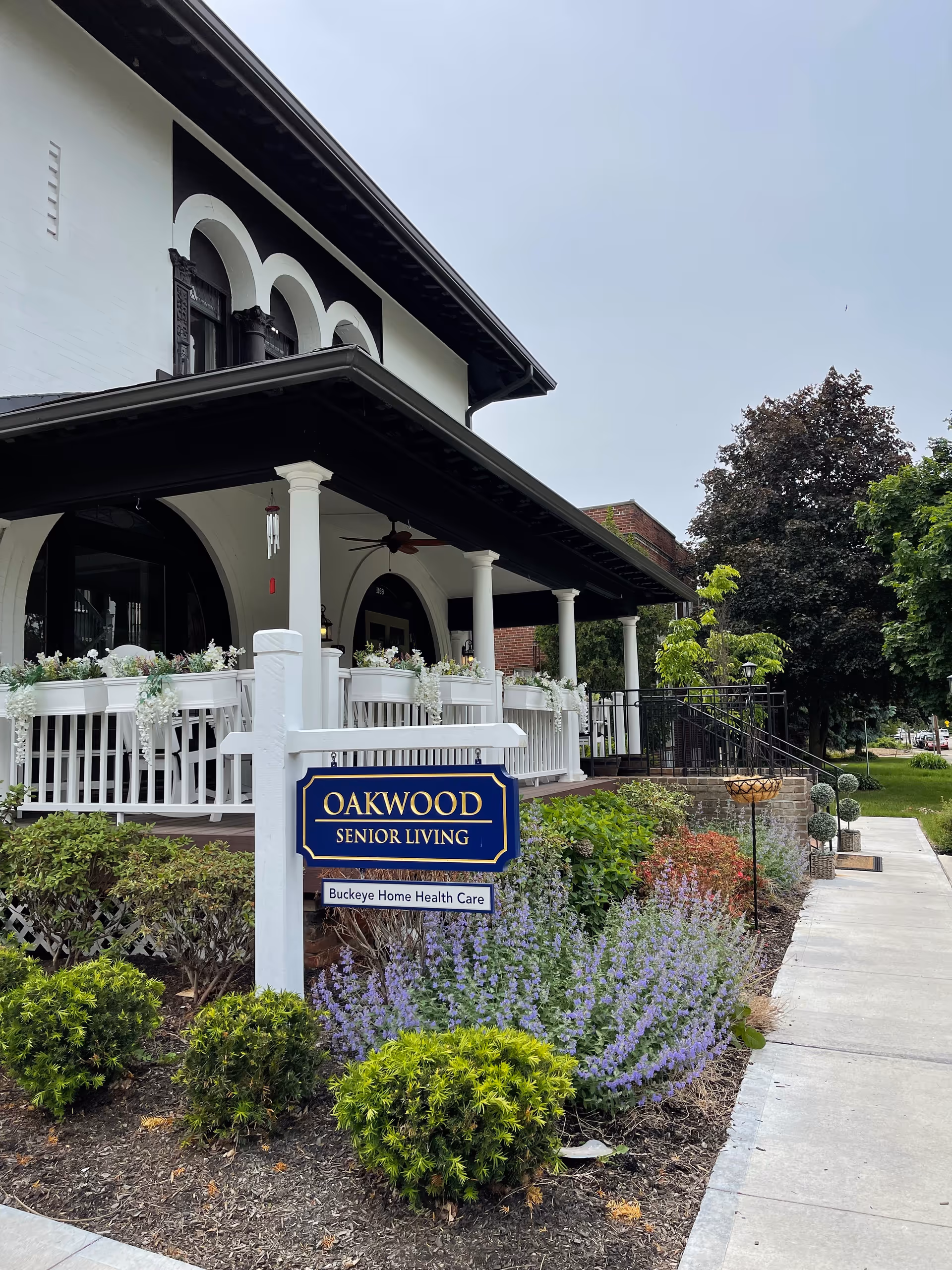 Exterior view of Oakwood Senior Living building with a porch featuring white railings and columns. There is a blue and gold sign in front that reads 'OAKWOOD SENIOR LIVING' with a smaller sign below it stating 'Buckeye Home Health Care'. The surrounding area has well-maintained landscaping with bushes, purple flowers, and a sidewalk leading past the building.
