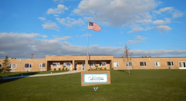 Front exterior view of Heritage of Emerson facility, a single-story brick building with multiple windows, a central entrance, an American flag on a flagpole, and a green lawn with a sign displaying the facility's name.
