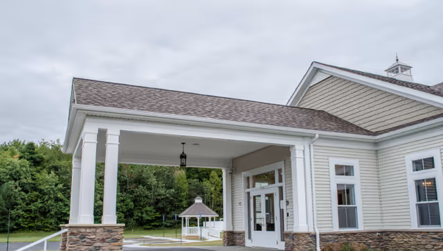 Covered porte-cochère entrance of a light-colored building with a small gazebo and wooded background.