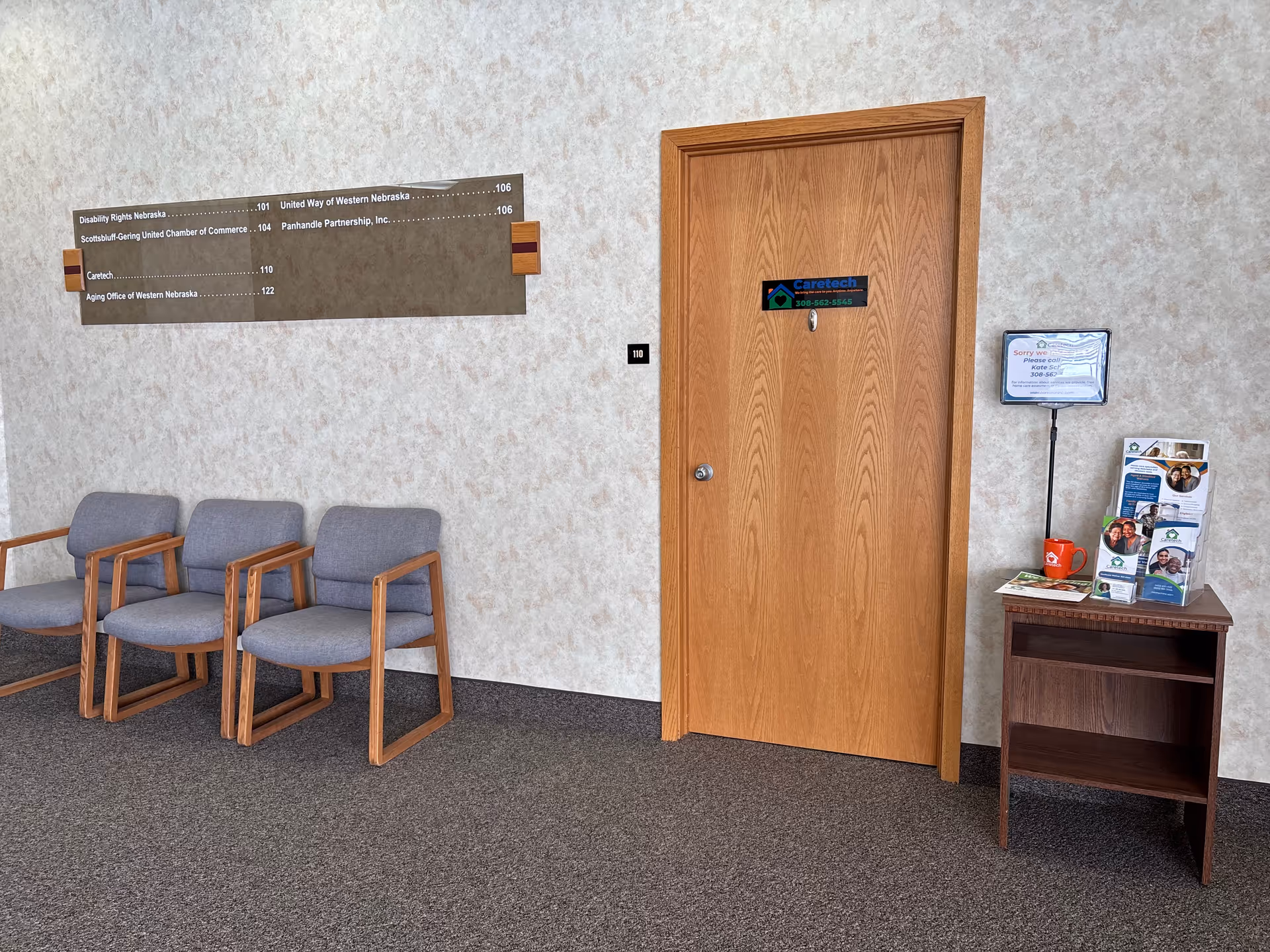 Interior hallway with a wooden door, seating area, and brochure stand in a senior facility.