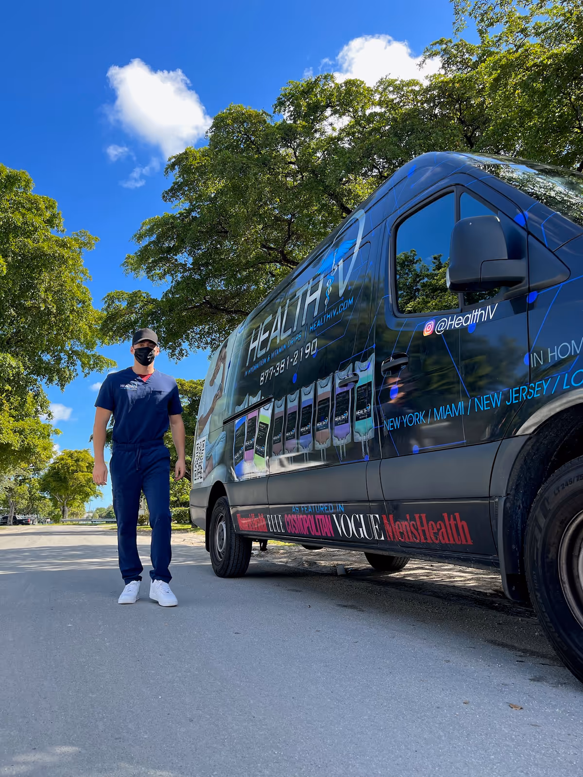 A man in scrubs stands beside a branded mobile IV therapy van on a tree-lined street.