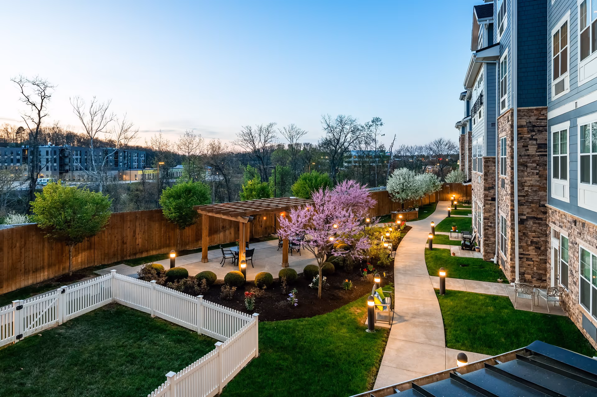 Well-lit landscaped courtyard with a winding walkway, pergola, flowering trees, and an adjacent multi-story building.