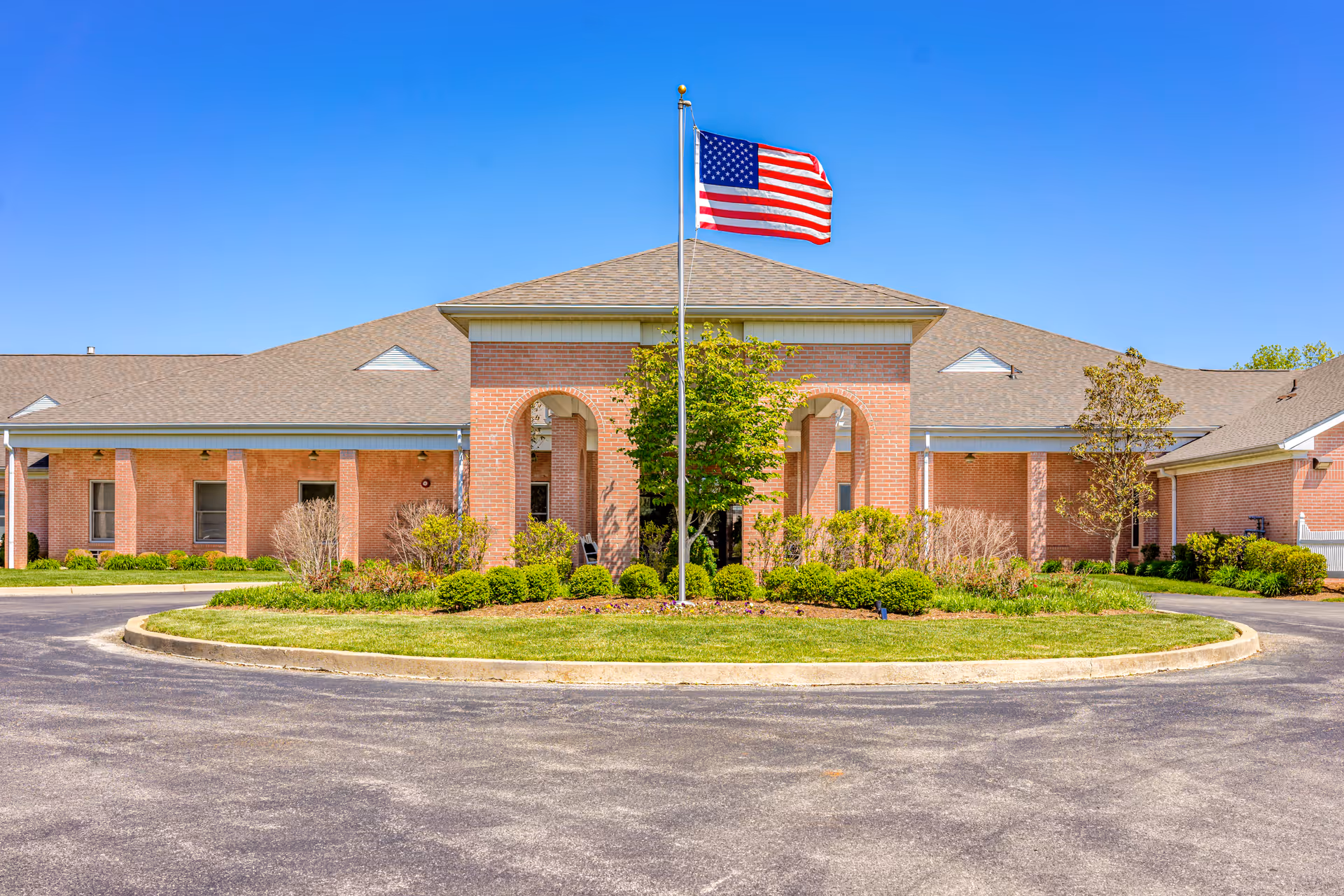 Front exterior view of a single-story brick building with a pitched roof, featuring a central entrance with arches and a flagpole flying the American flag in front. The building is surrounded by landscaped greenery and a circular driveway under a clear blue sky.