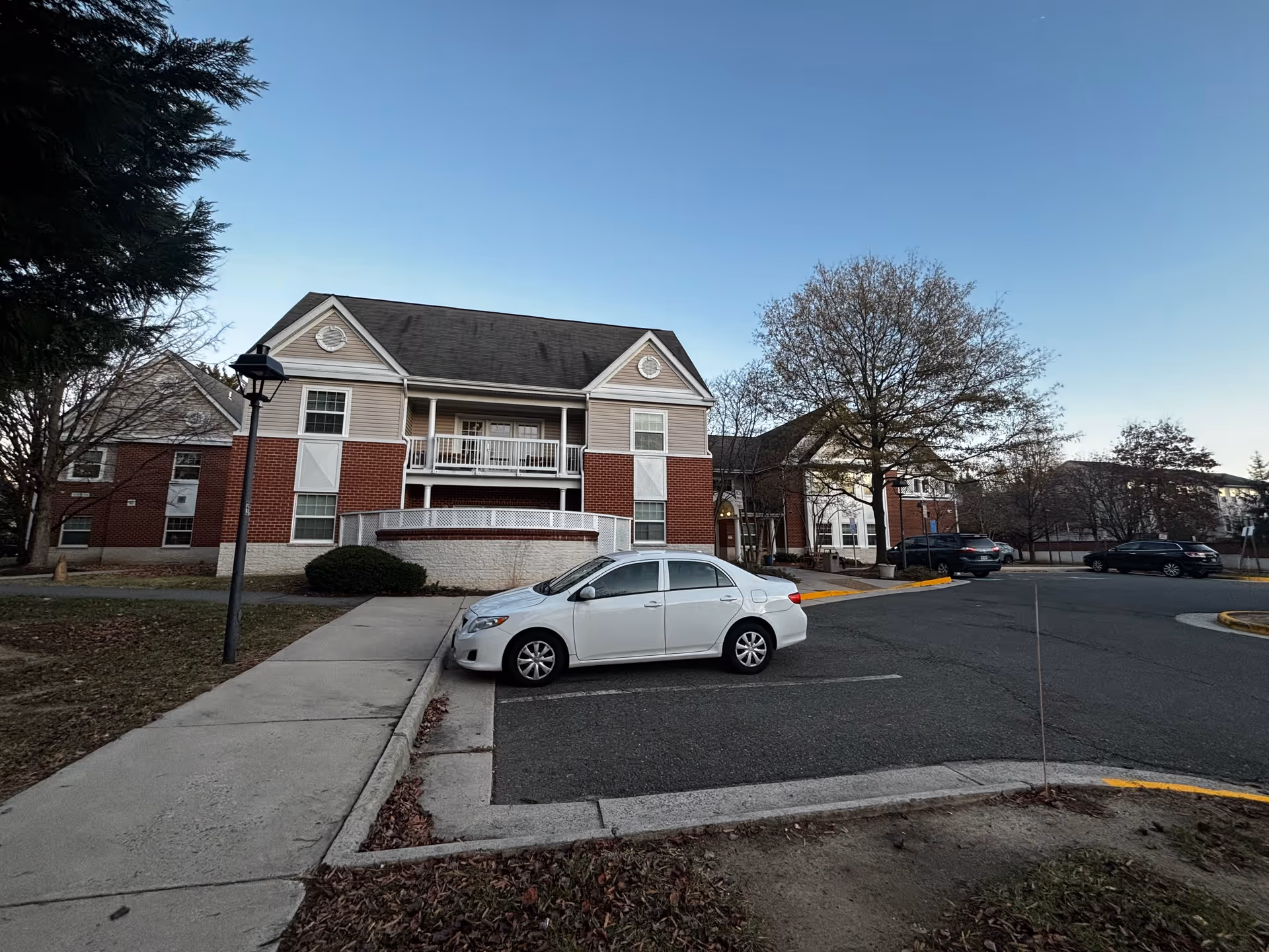 Front view of a two-story brick-and-siding senior living building with parked cars, a large leafless tree, and a sidewalk.
