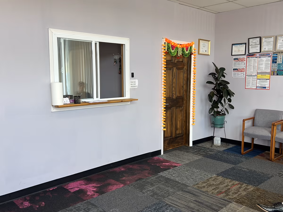 Interior reception area featuring a service window, decorated door with garland, plant, and chairs.