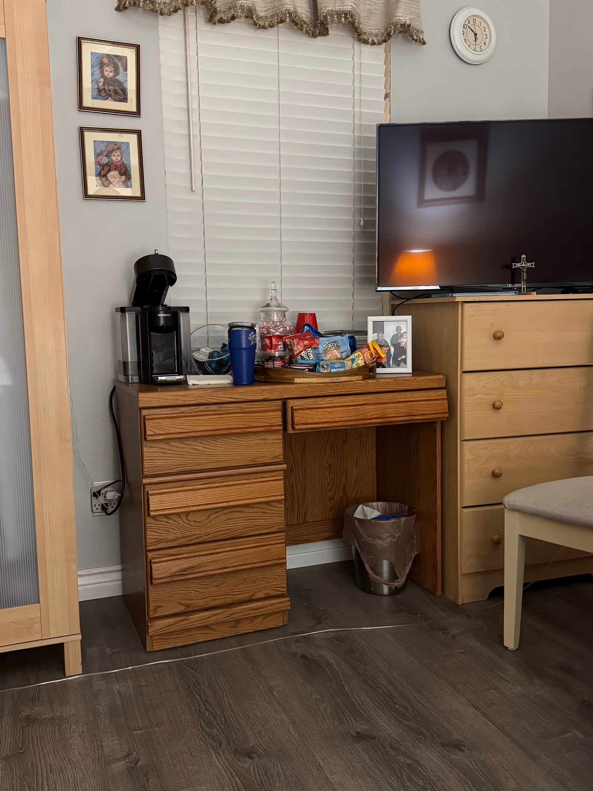Wood desk with coffee maker, snacks, photos, and a TV across from a chair.