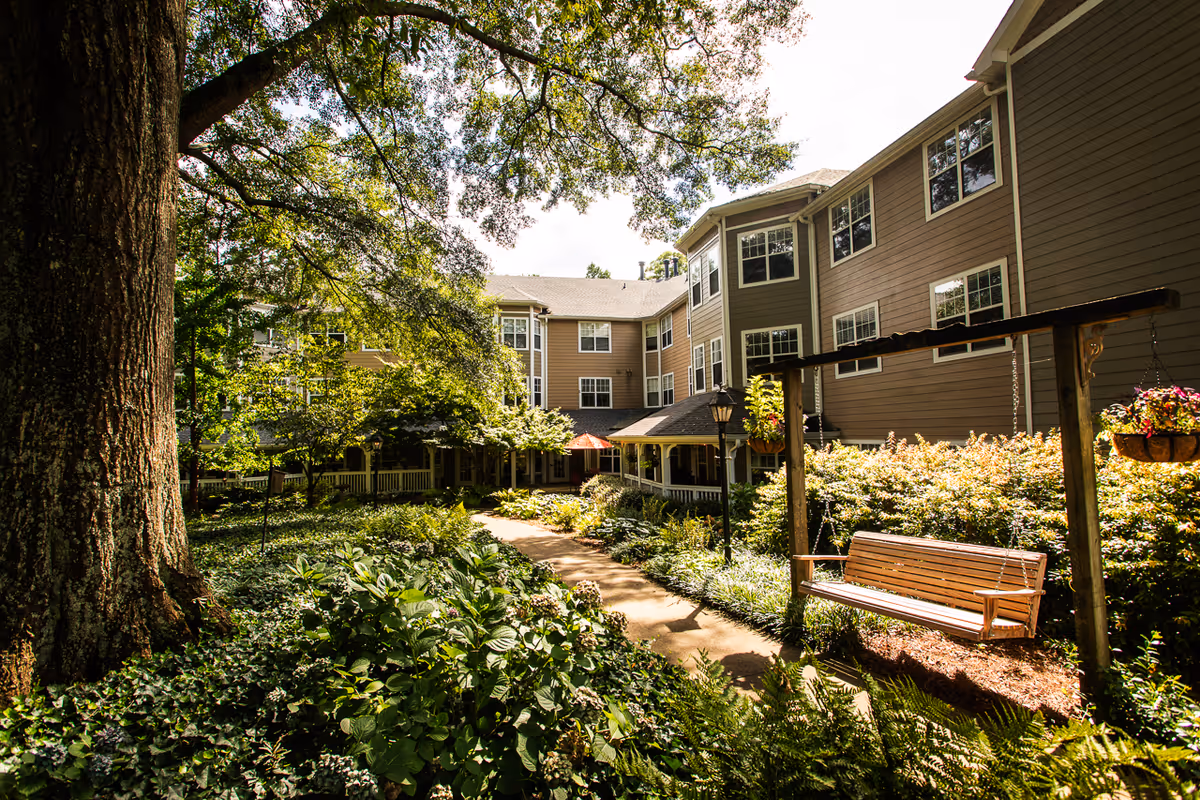 A peaceful outdoor garden area at Winnwood Retirement Community featuring a large tree, lush greenery, a paved walking path, and a wooden swing bench. The multi-story building with many windows surrounds the garden, and hanging flower baskets add color to the scene.