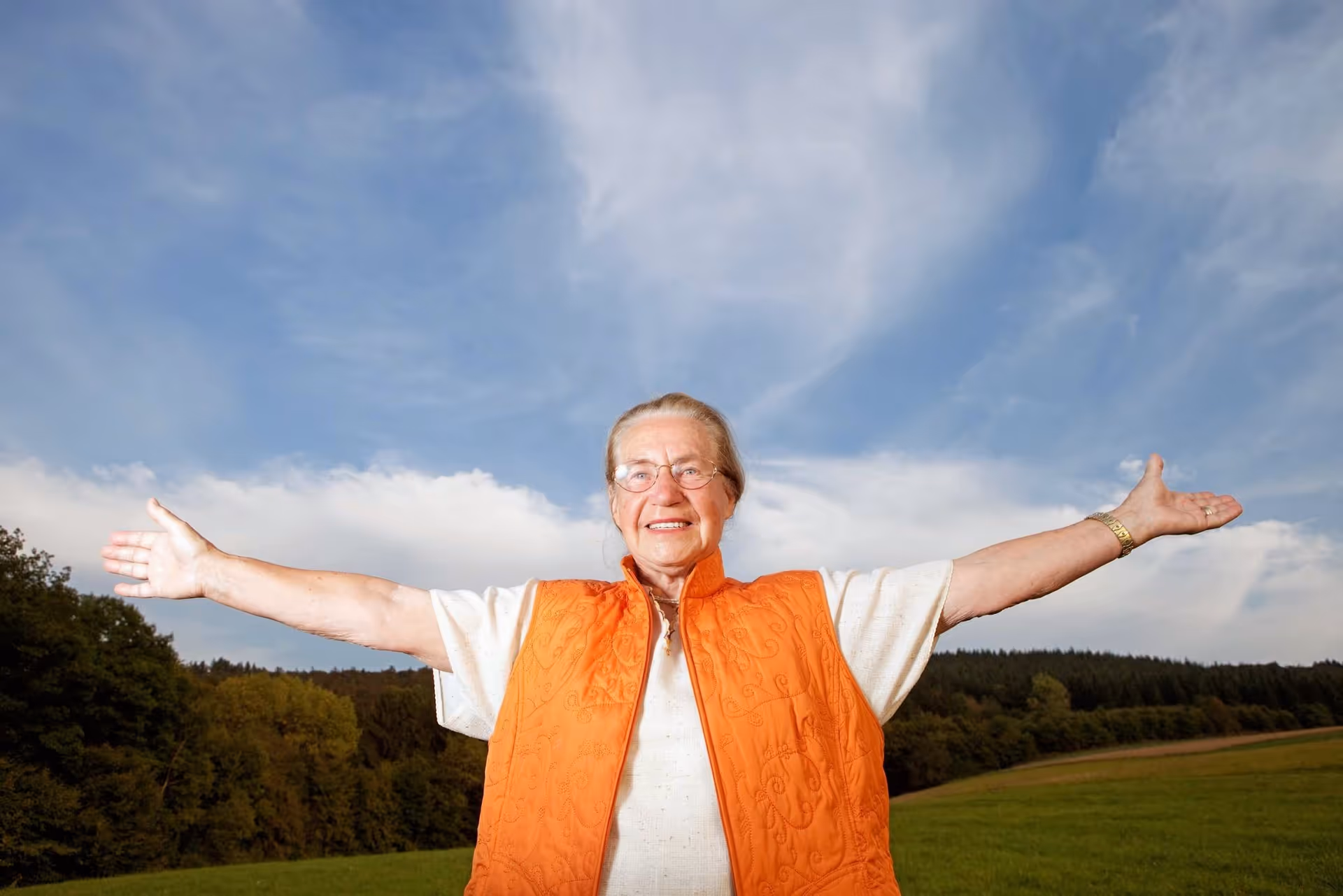 Senior woman outside with arms wide, smiling, wearing an orange vest under a blue sky.