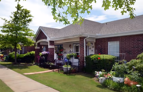 Exterior view of a single-story brick building with a covered porch, potted plants, and a small garden area. A sidewalk runs alongside the building, and there are trees and shrubs in the landscaped yard under a partly cloudy sky.