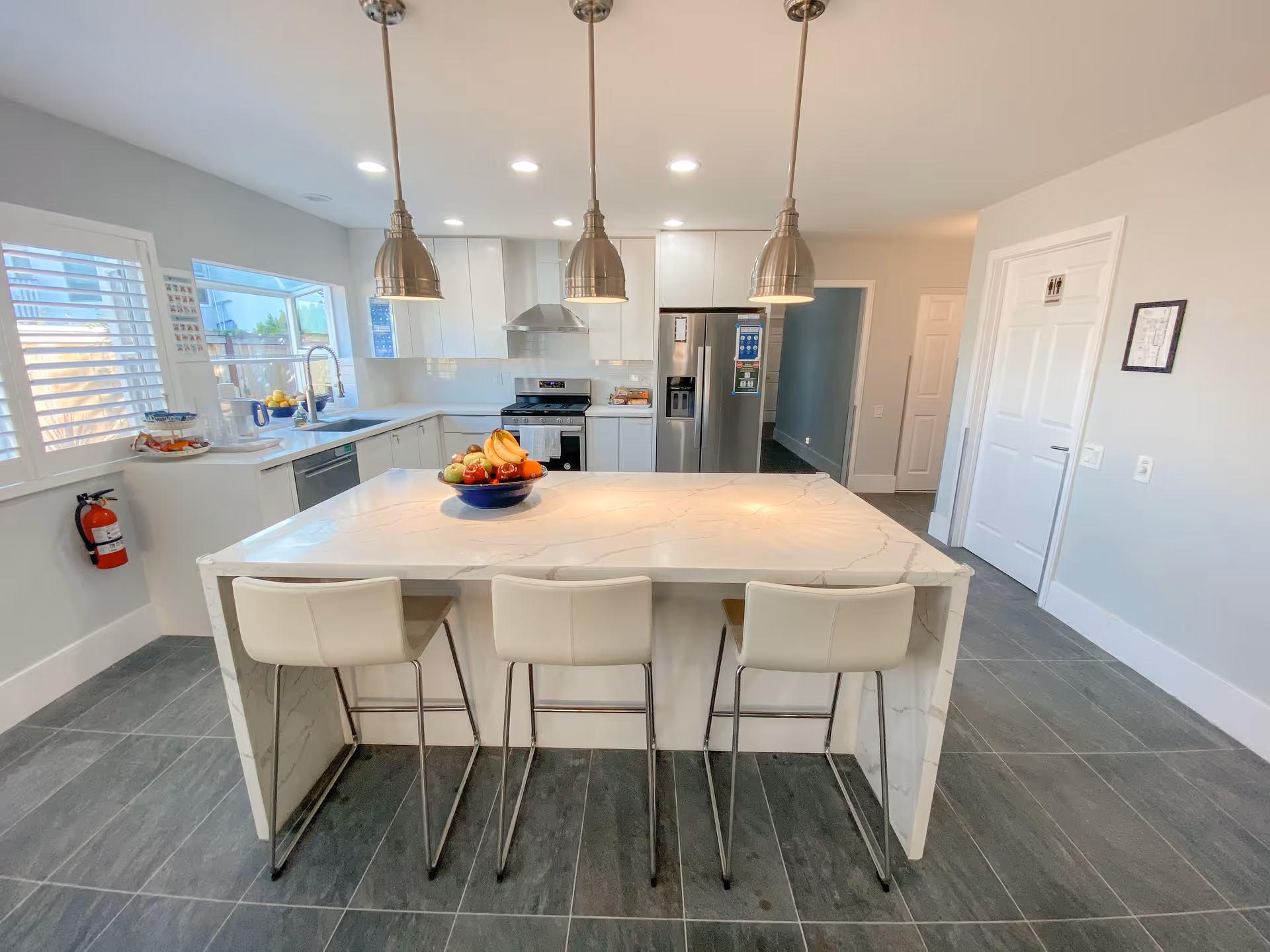 Bright modern kitchen with marble island, three stools, stainless appliances, and large windows.