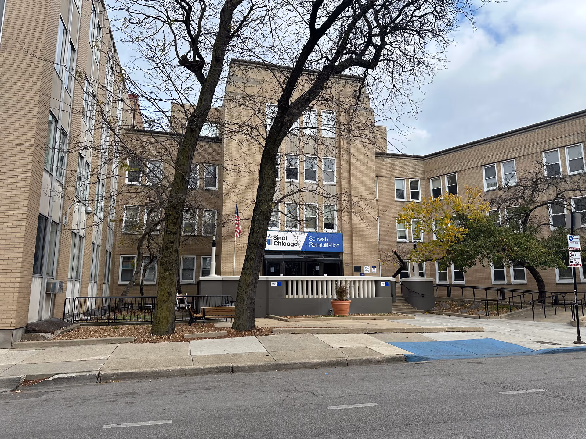 Exterior view of Schwab Rehabilitation Center SNU, a multi-story beige brick building with numerous windows. There are leafless trees and some greenery in front of the building, along with a sidewalk and a street. A sign above the entrance reads 'Sinai Chicago Schwab Rehabilitation.'
