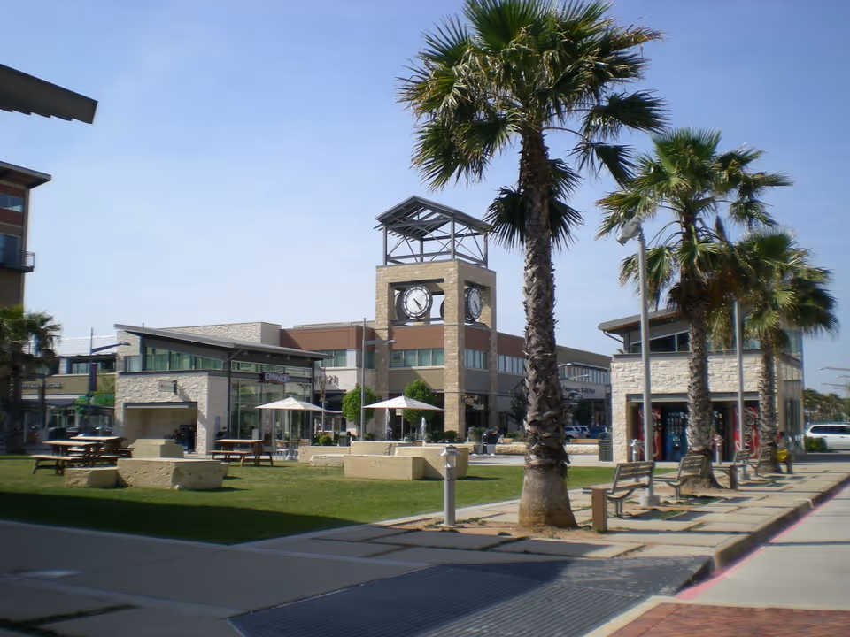 Outdoor shopping plaza with palm trees, benches, and a central clock tower.