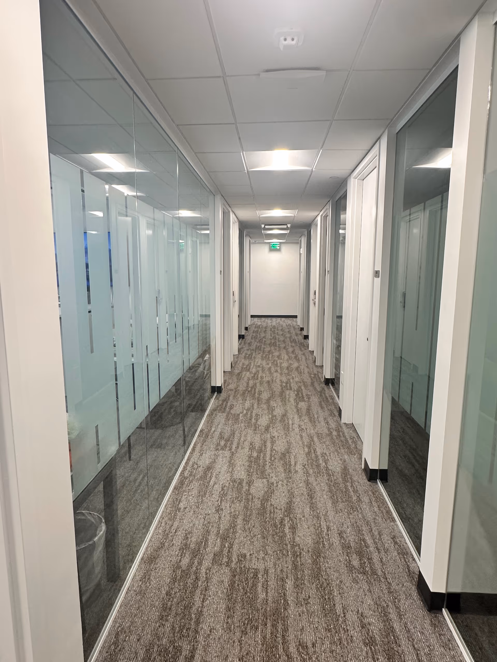 A long, glass-walled hallway with doors on both sides and bright ceiling lights.