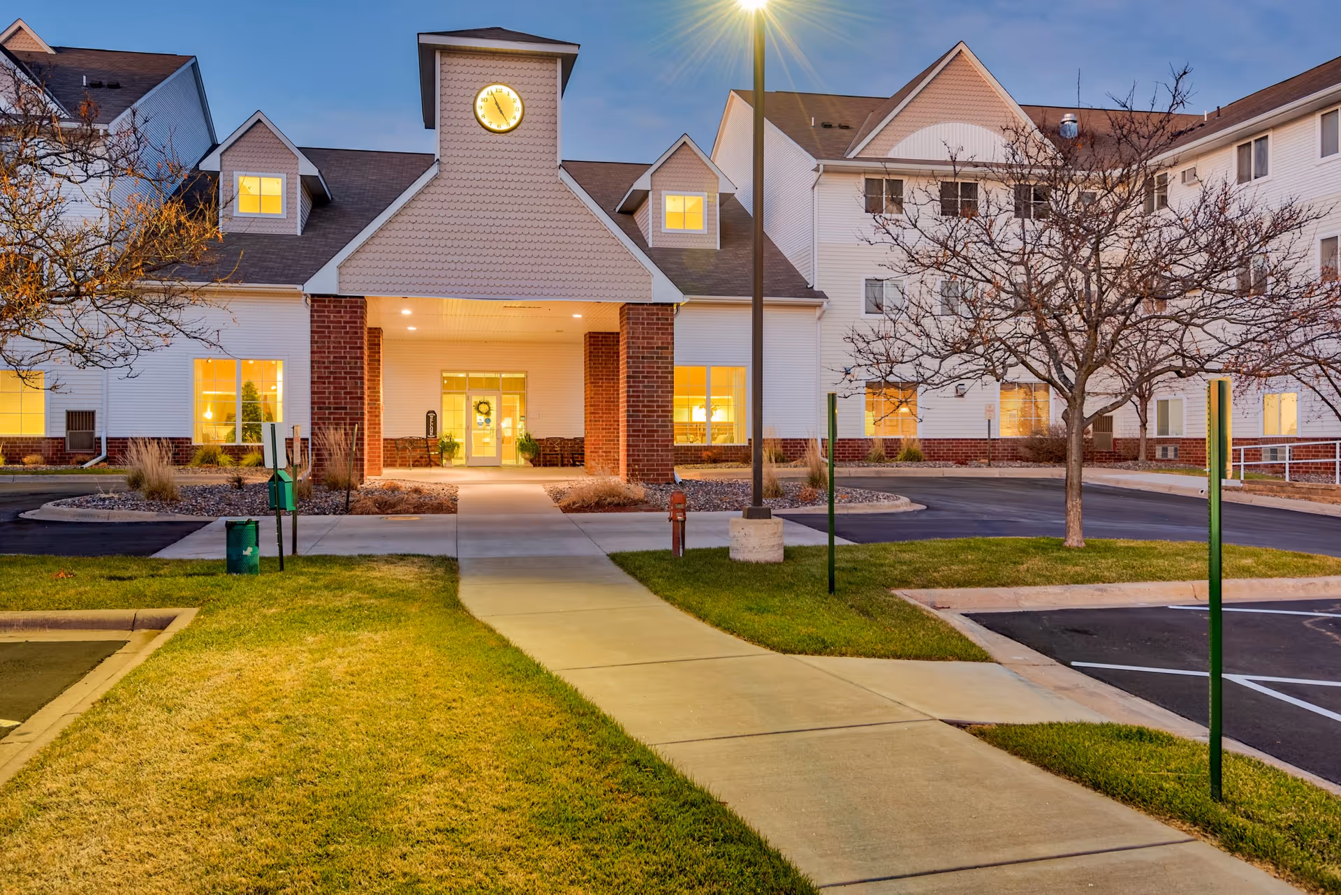 Exterior front entrance of a senior living facility at dusk with a clock tower, illuminated windows, paved walkway, and lawn.