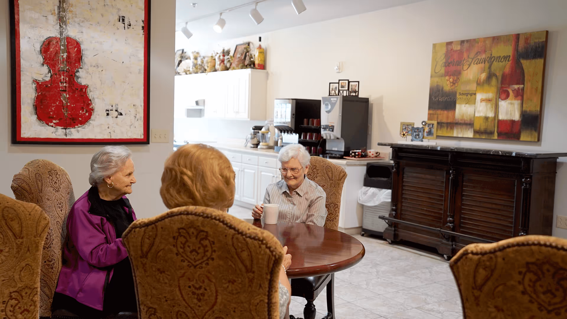 Three elderly women sit and chat around a round wooden table in a well-lit common dining area with artwork and a coffee station.