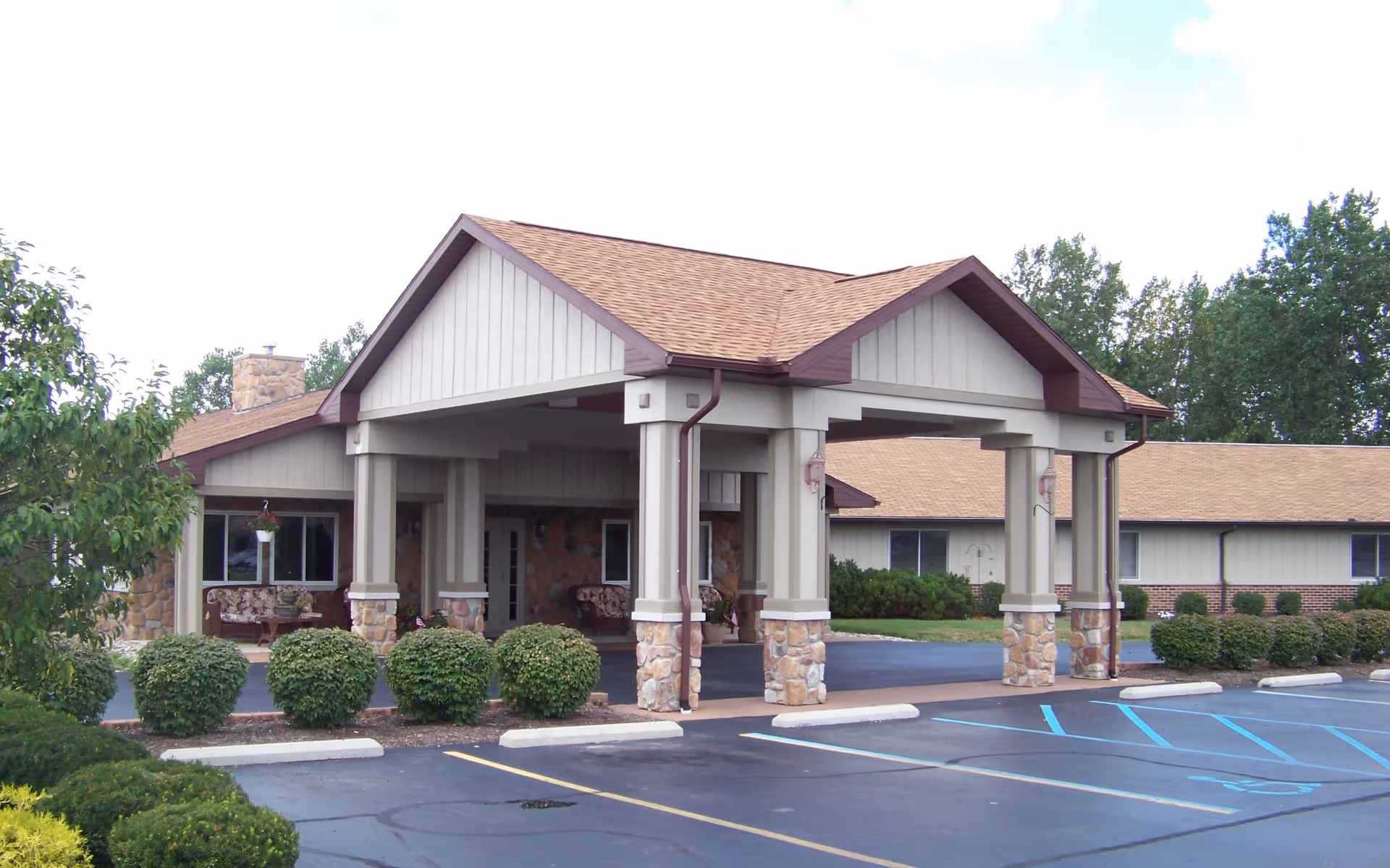 Exterior view of Briar Hill Health Campus - Memory Care showing the front entrance with a covered drop-off area supported by stone and beige pillars. The building has a tan roof and beige siding with stone accents. There are neatly trimmed bushes and a parking lot with marked spaces, including handicapped parking spots.