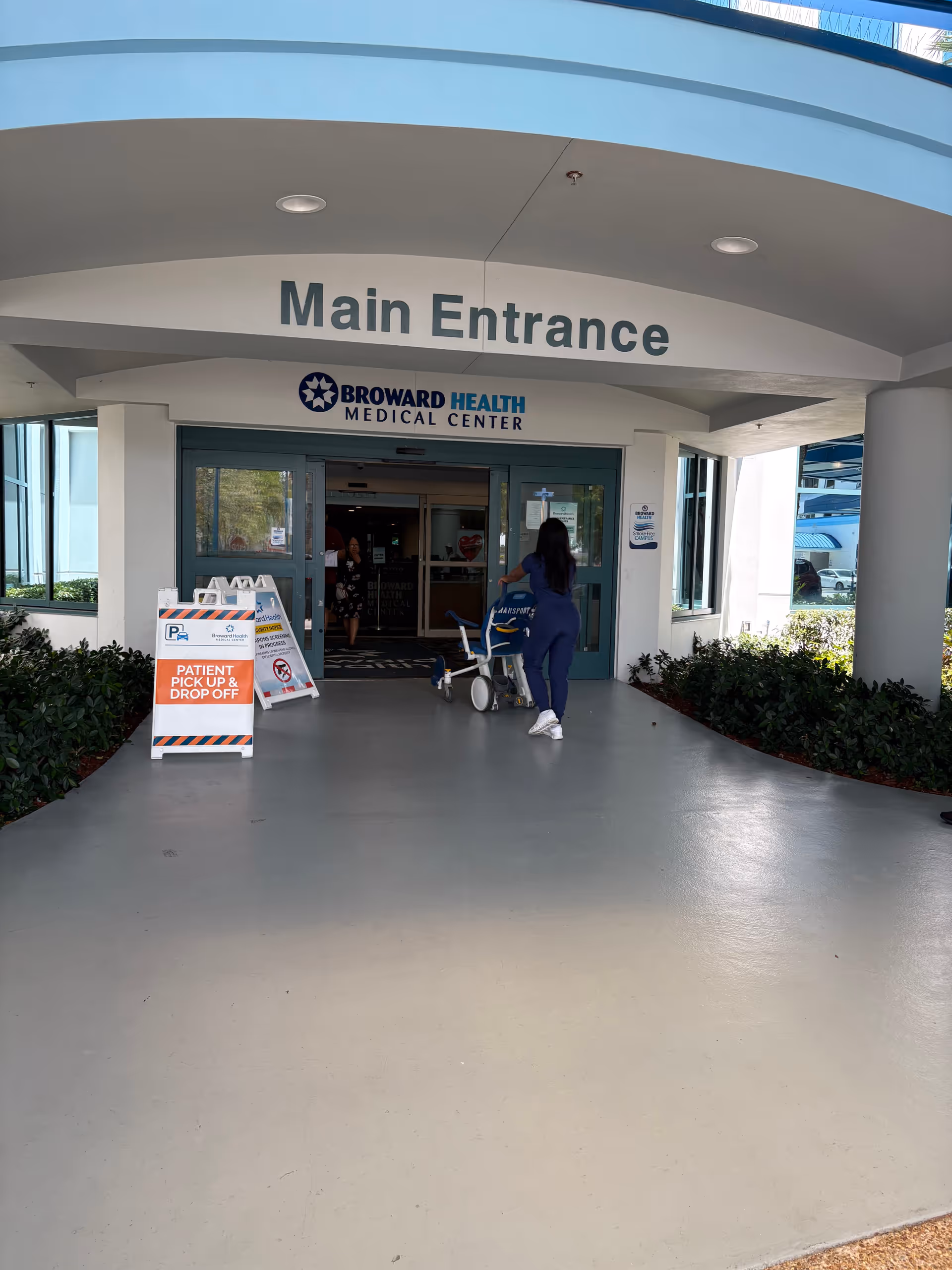 Main entrance of a hospital with a person pushing a cart toward glass doors.