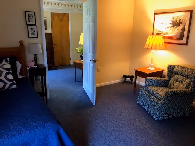 Bedroom in a senior living suite with a blue bedspread, seating, and lamps.
