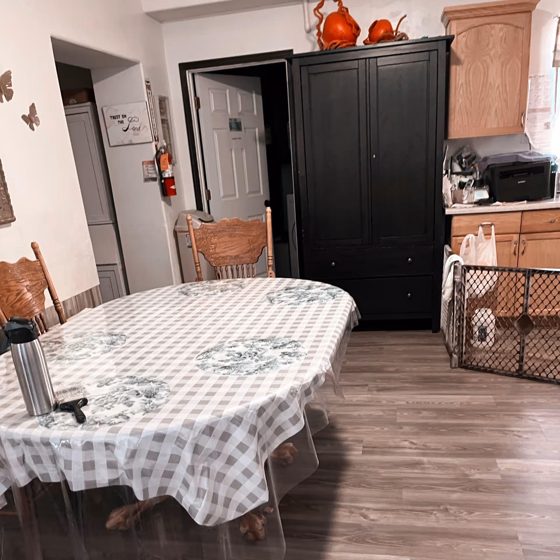 Interior view of a dining area with a wooden table covered by a checkered tablecloth with floral patterns. There are wooden chairs around the table. In the background, there is a black cabinet with decorative items on top, wooden kitchen cabinets, a printer, and a baby gate. The floor is wood laminate.