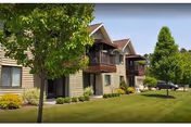 Two-story senior living building with balconies, a landscaped lawn, trees, and a parked car.