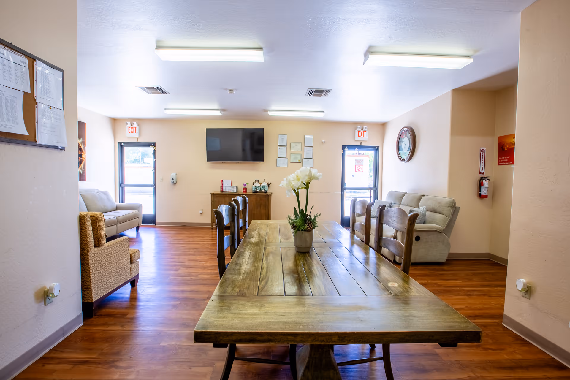 A spacious senior living common area featuring a long wooden dining table and surrounding seating.