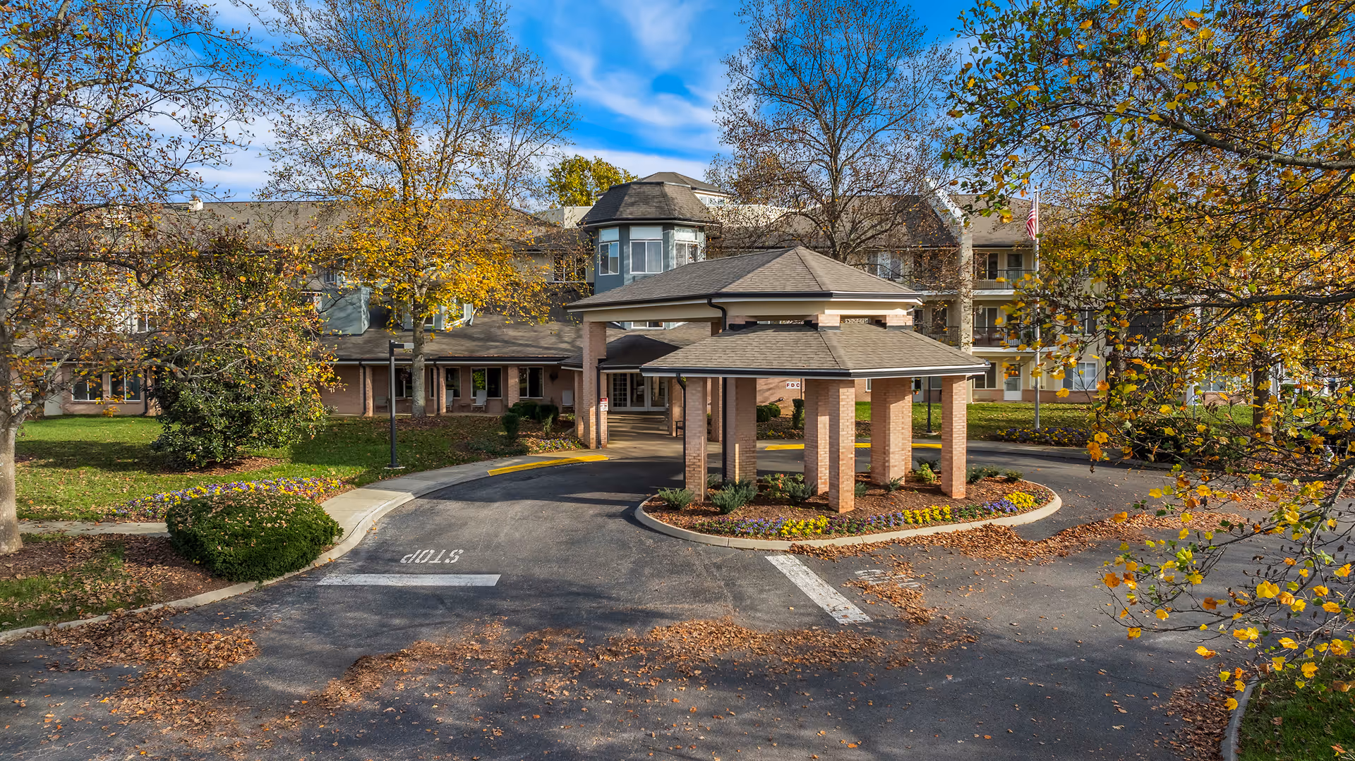 Exterior view of a senior living community building with a covered circular driveway entrance surrounded by trees with autumn foliage and landscaped flower beds.