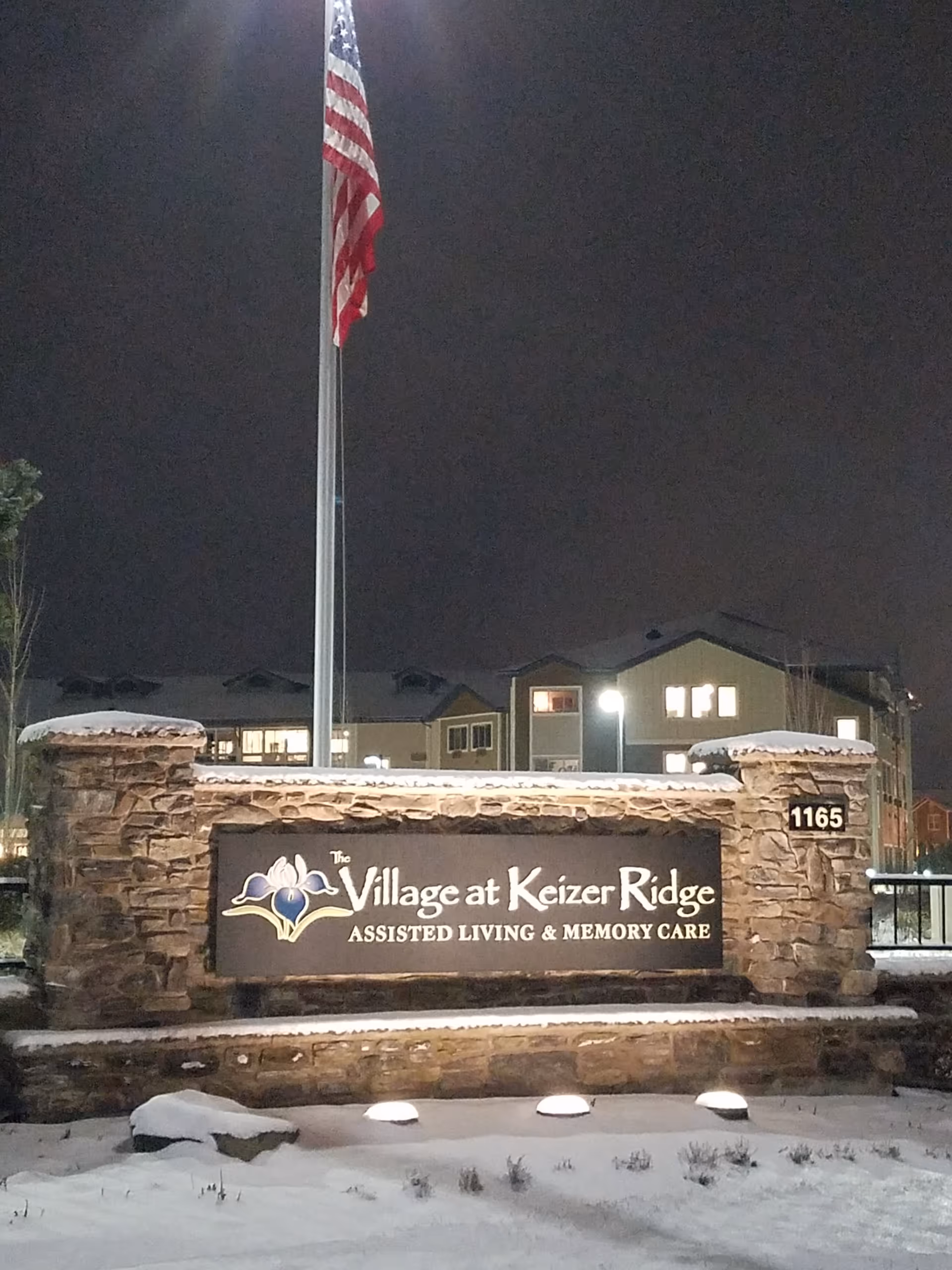 Stone entrance sign reads The Village at Keizer Ridge Memory Care with American flag and snow.