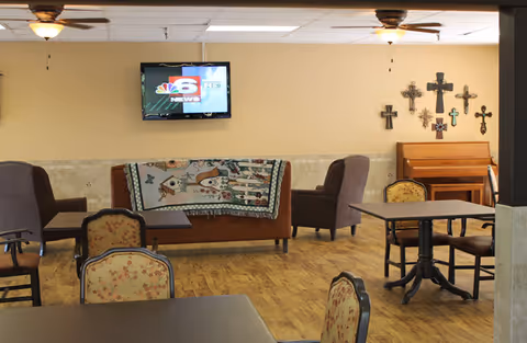Senior living common room with seating, a TV, a piano, and wall crosses.