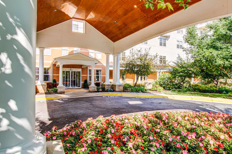 Covered circular driveway and main entrance of a brick senior living building framed by columns, landscaping, and flower beds.