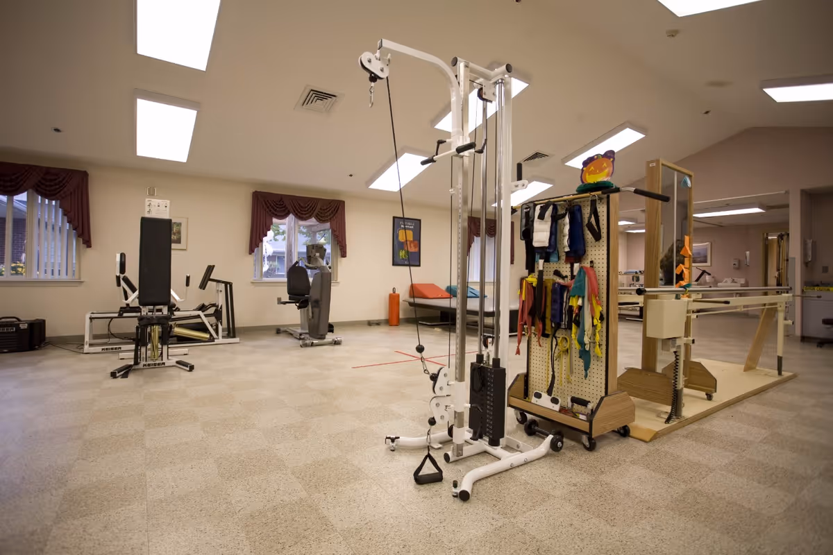 A spacious rehabilitation or physical therapy room with exercise equipment including a weight machine, stationary bike, and parallel bars. The room has beige walls, windows with maroon curtains, and bright overhead lighting.