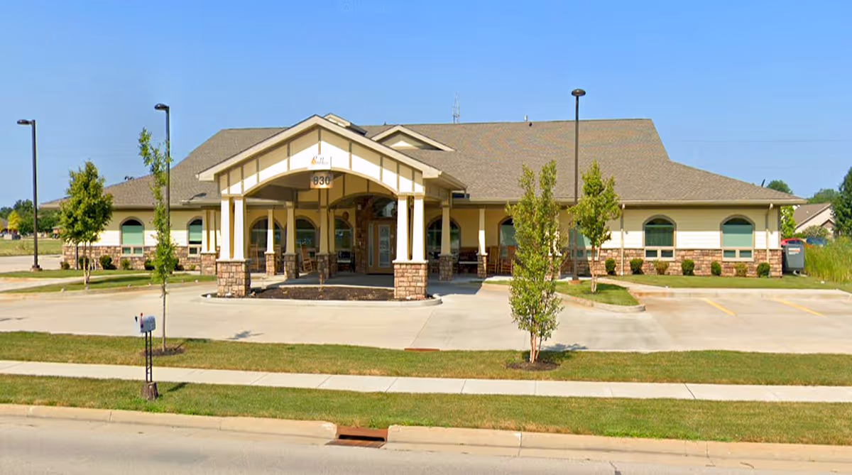 Single-story memory care building with a covered entrance, parking area and small trees on a sunny day.