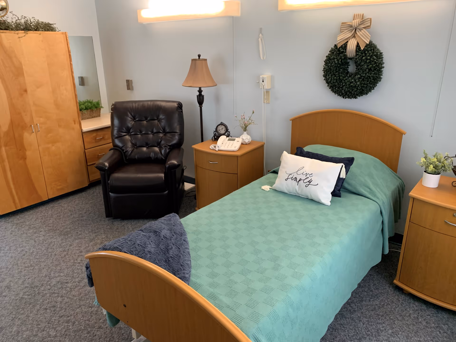A neatly arranged bedroom in a care facility featuring a single bed with a green bedspread and decorative pillows, including one that says 'live simply'. There is a wooden headboard and matching wooden nightstands on either side of the bed. A black leather recliner chair is positioned next to a floor lamp and a small table with a telephone and clock. A wooden wardrobe and dresser with a mirror are visible in the background. A green wreath with a striped bow hangs on the wall above the bed.