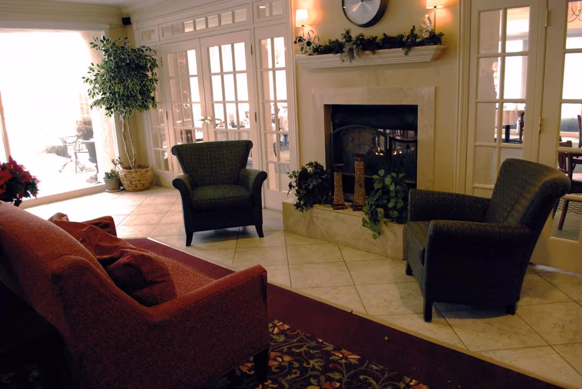 A cozy living room area with a lit fireplace, two dark upholstered armchairs, and a red sofa. The room has tiled flooring and a patterned rug. There are glass French doors behind the armchairs, and a large potted plant is placed near the doors. The fireplace mantel is decorated with greenery and a large clock above it.
