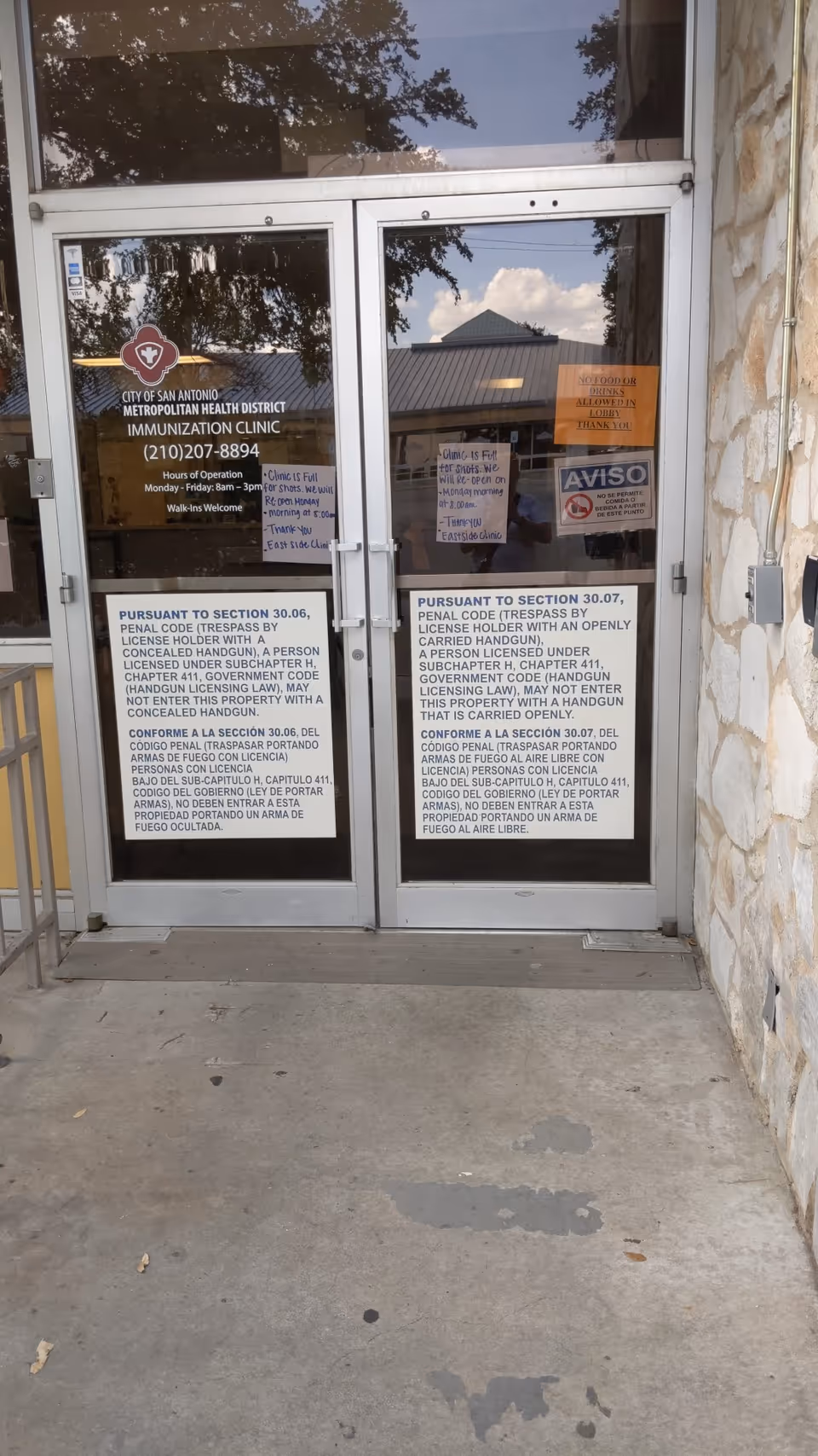 Double glass doors with posted notices and reflected trees at clinic entrance.