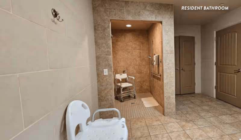 A spacious resident bathroom with beige tiled walls and floor. The image shows a walk-in shower area with a white shower chair inside. There is a white bath chair with armrests positioned outside the shower. Two closed brown doors are visible in the background. The words 'RESIDENT BATHROOM' are written in white text in the upper right corner.