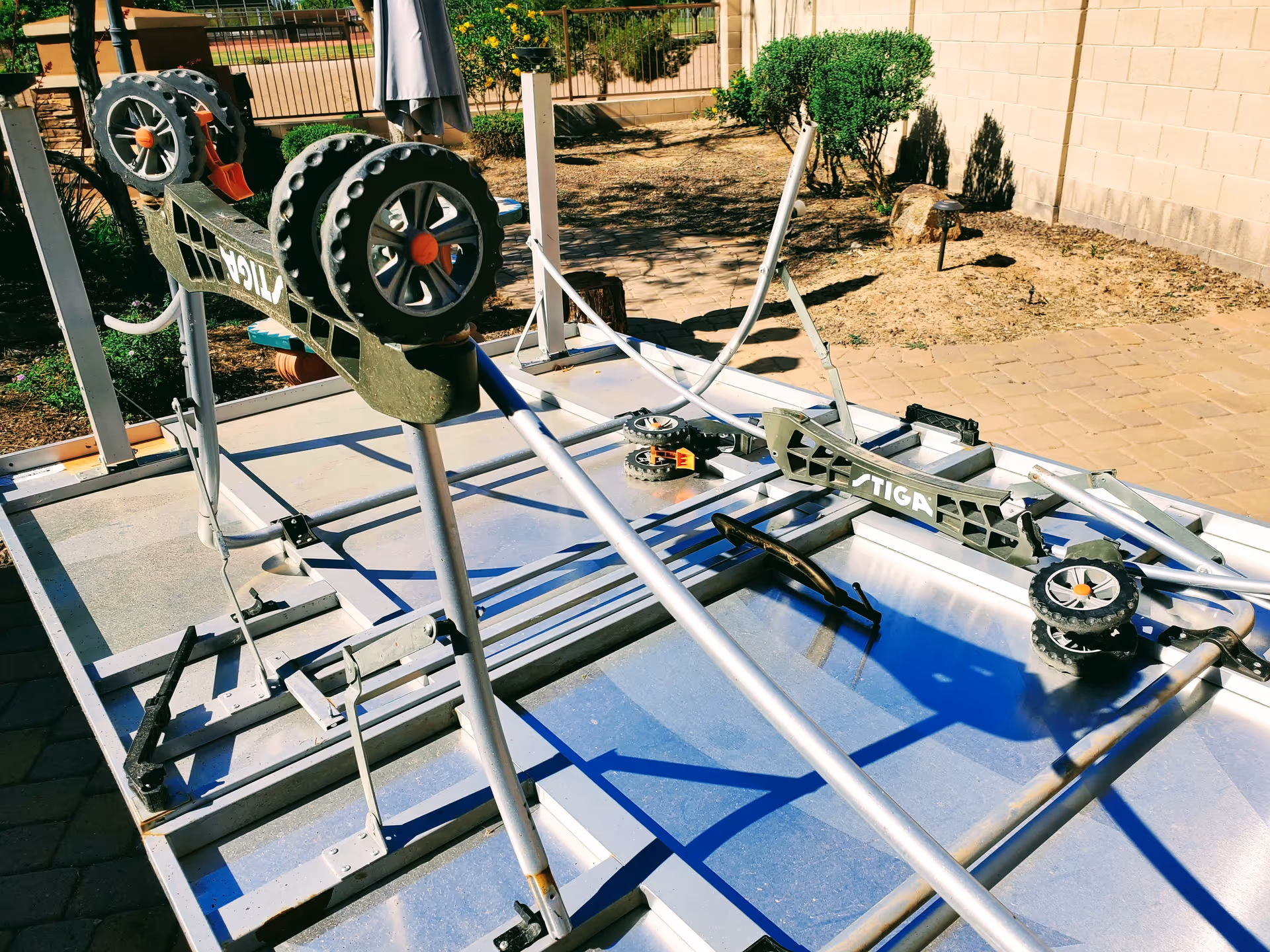 Outdoor metal exercise equipment on a blue platform beside a brick wall and bushes.