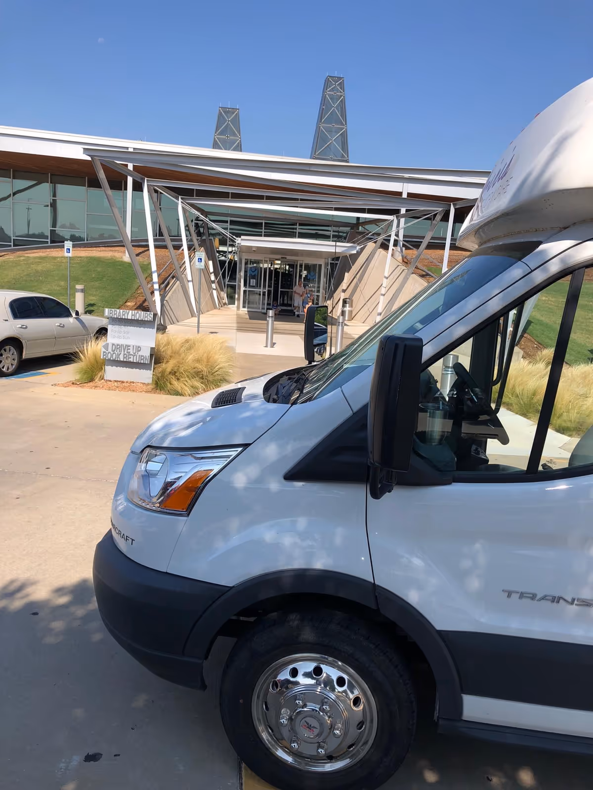 Front exterior view of a modern building with large glass windows and a covered entrance supported by metal beams. A white van is parked in the foreground on the right side, and a silver car is parked on the left. There is a sign near the entrance indicating library hours and a drive-up book return. The sky is clear and blue.