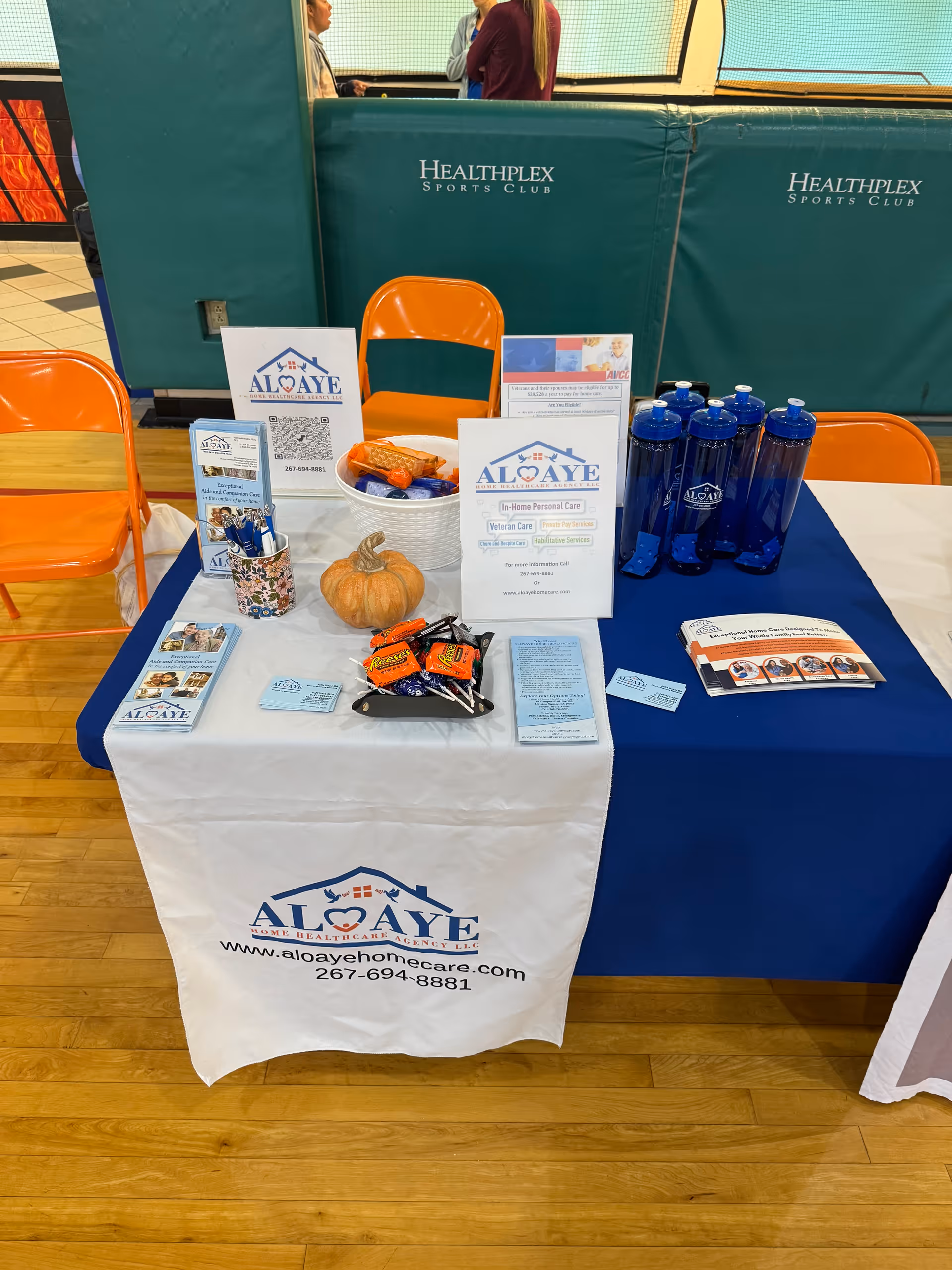 Promotional booth for Aloaye Home Care Agency with brochures, pumpkins, and blue water bottles at gym