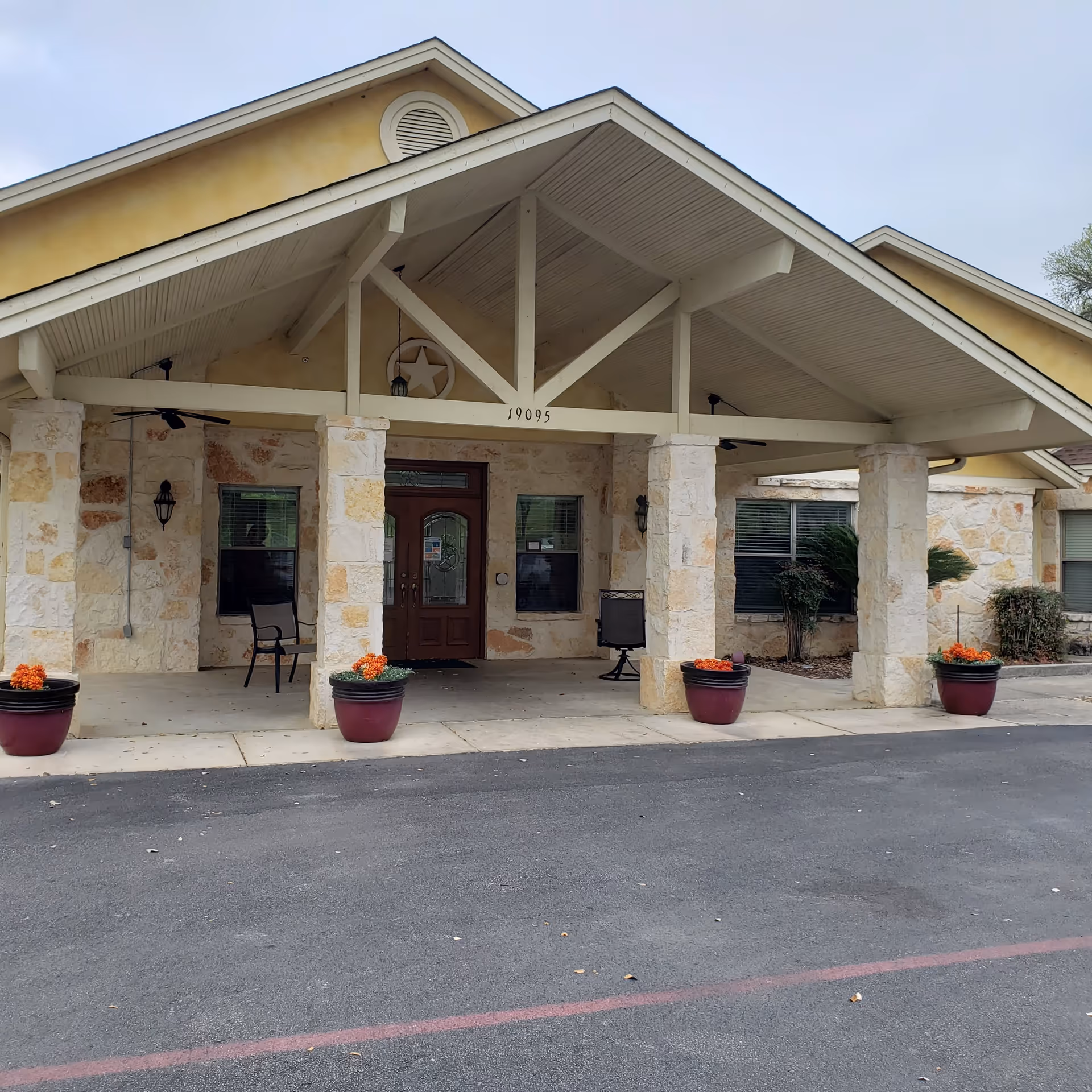 Front exterior view of a building with stone pillars and a covered entrance. There are four large flower pots with orange flowers placed symmetrically in front of the entrance. The building has a yellow upper facade and a brown double door with glass panels. The address number 19095 is displayed above the entrance.