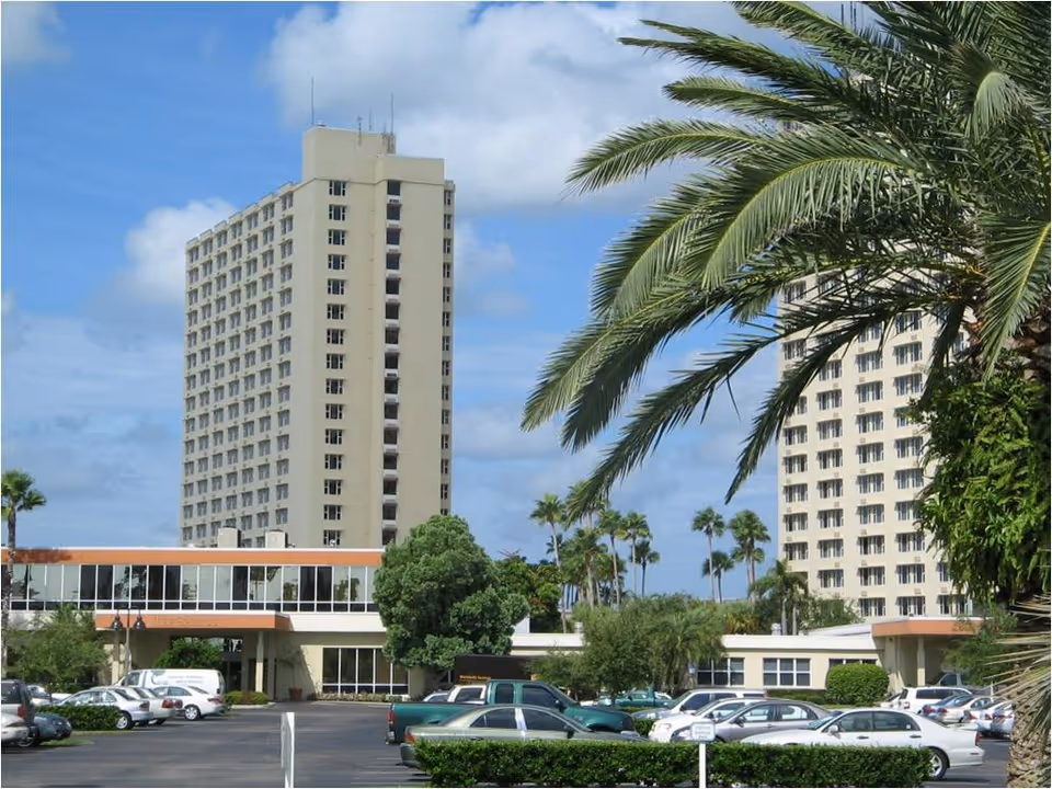 Two tall beige residential towers with a parking lot and palm trees in foreground.