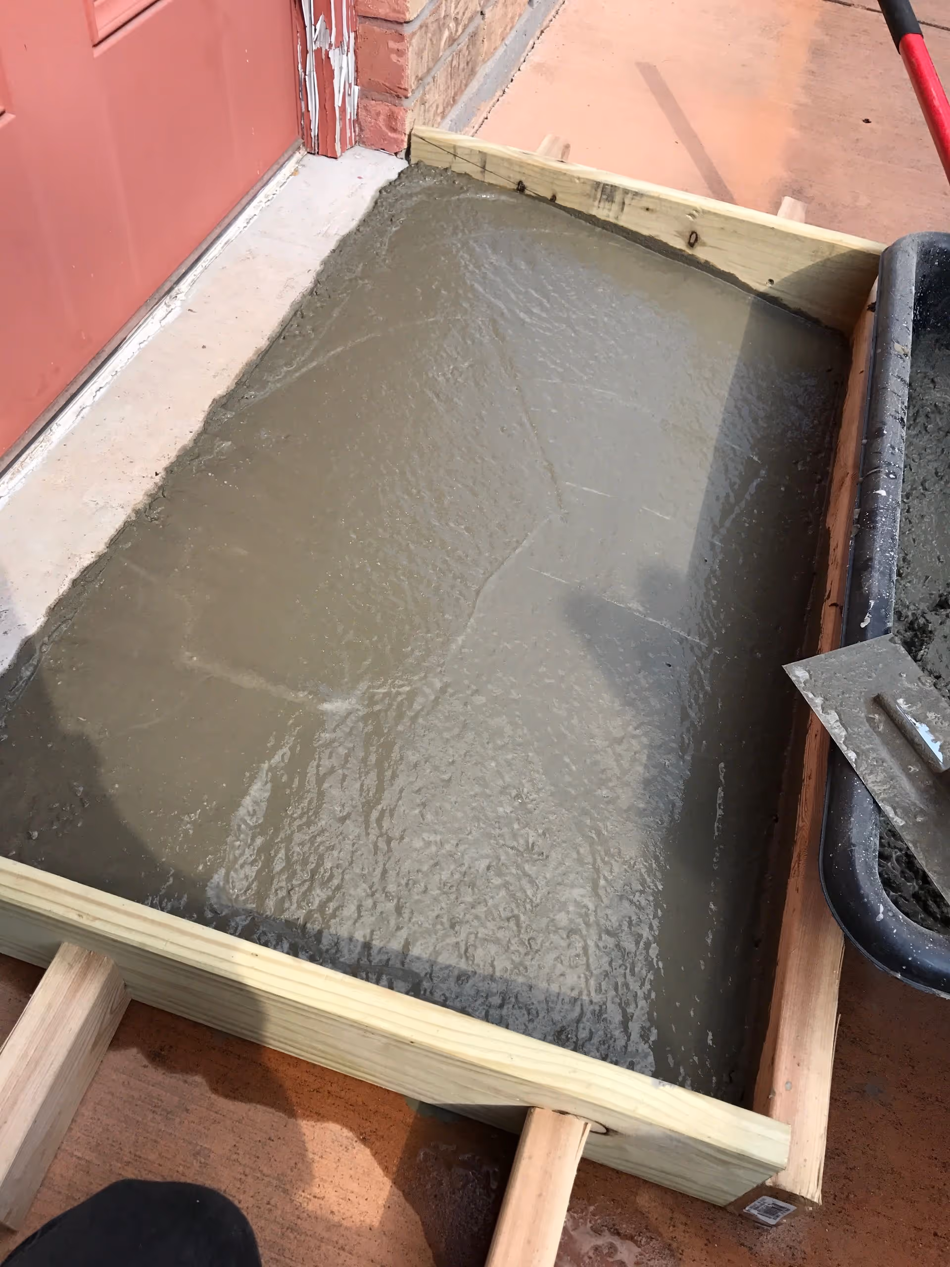 Freshly poured concrete in a wooden form beside a red door outdoors.