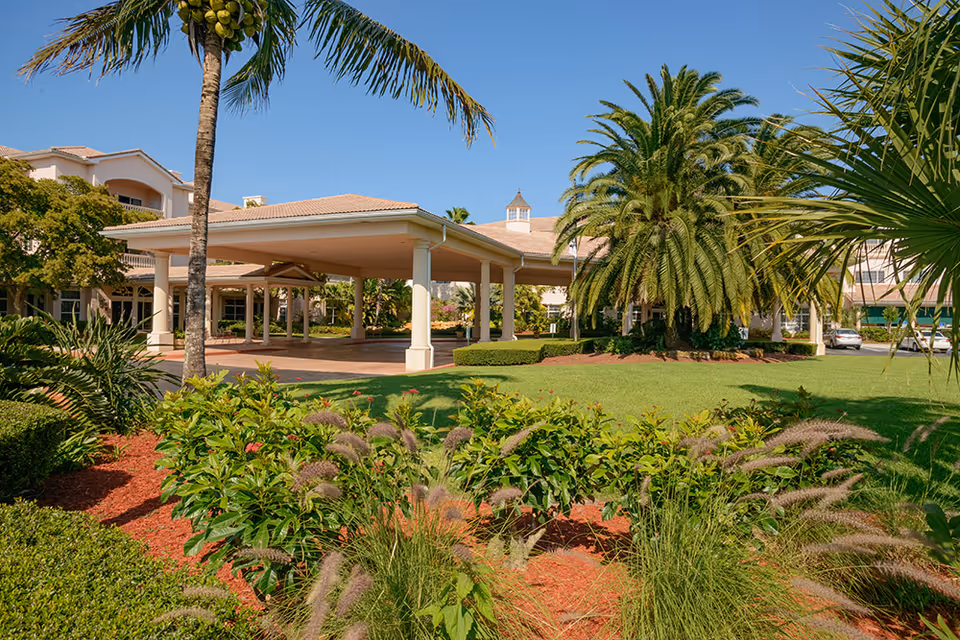 Exterior view of Harbor Place at Port St. Lucie featuring a covered entrance with columns, surrounded by lush landscaping including palm trees, bushes, and green grass under a clear blue sky.