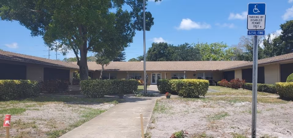 Single-story building with a tan exterior and a brown roof, surrounded by trimmed bushes and trees under a blue sky with some clouds. A concrete walkway leads to the entrance doors in the center. A signpost near the walkway indicates parking is reserved for disabled permit holders with a $250 fine for violations.