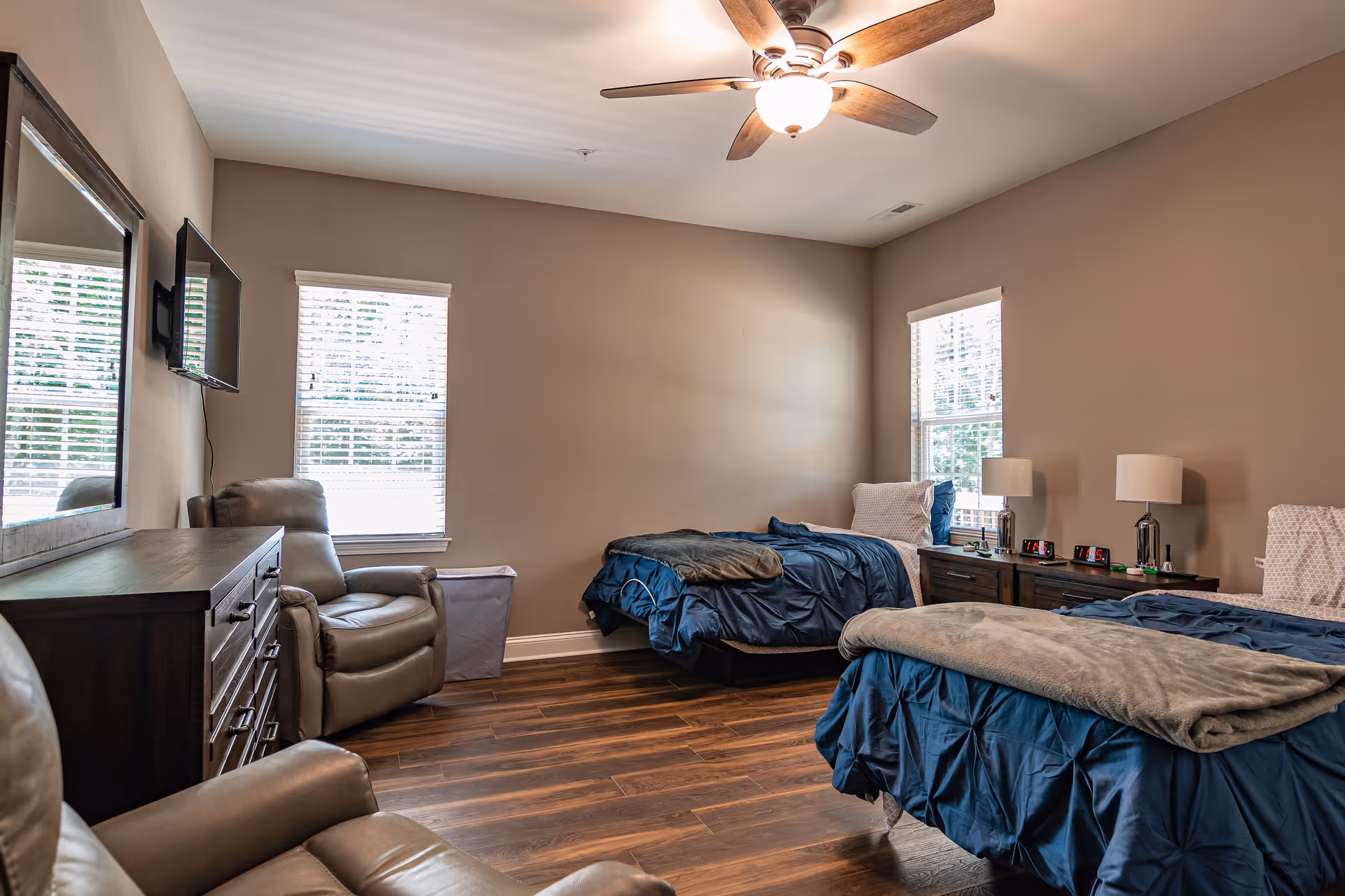 A shared senior living bedroom with two beds, recliners, dresser, and a wall TV.