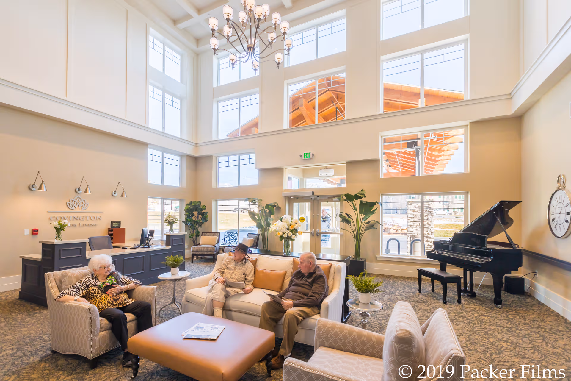 Bright, open senior living lobby with sofas, a grand piano, reception desk, and large windows.